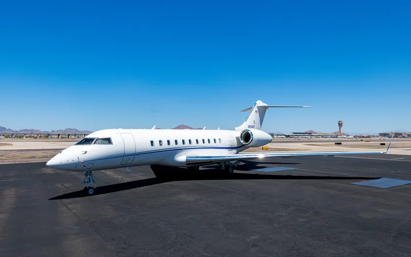 Jet privado Bombardier Global 5500 aparcado en la plataforma bajo un cielo azul despejado, con la torre de control del aeropuerto y montañas al fondo