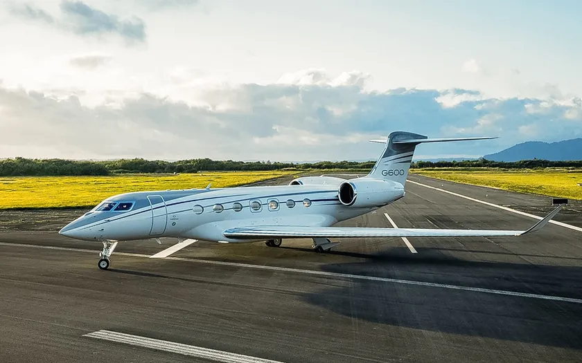 Jet privado Gulfstream G600 en una pista con campos verdes y montañas al fondo