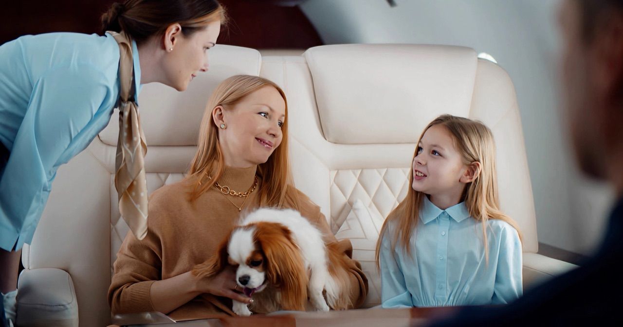A mother cradling a small dog beside her young daughter in a private jet cabin, welcomed by a smiling flight attendant