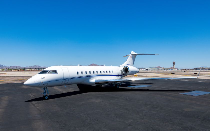 Bombardier Global 5500 private jet parked on the apron under a clear blue sky, with an airport control tower and mountains behind